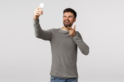 Cheeky, excited and carefree young man bearded, travel abroad photographing, posting pictures online, use application filters, hold smartphone upwards and taking selfie, wink, white background.