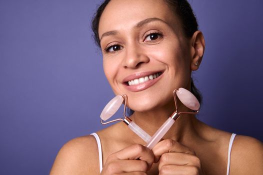 Close-up portrait of a natural beauty middle aged attractive African woman, smiling with cheerful toothy smile holding two pink jade roller stone massager in her hands, isolated on violet background