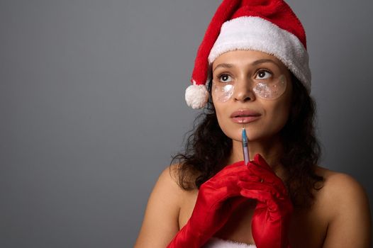 Pretty woman with natural make-up and patches under eyes, in Santa hat holds syringe with beauty injection near her face, looks up on copy space of gray background. Injection cosmetology, spa concept