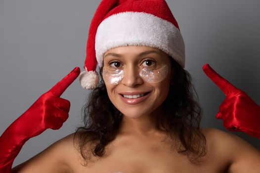 Attractive young woman in Santa costume points fingers on eye patches on her face, smiles looking at camera. Advertising for beauty salons for Christmas and New year giveaways. Spa, skin care concept