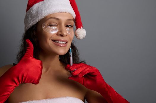 Attractive woman with under eyes patches, dressed in Santa , holds syringe with beauty injection, shows thumb up, smiles to camera. Anti-aging, face lifting, rejuvenation, cosmetology concept for ad