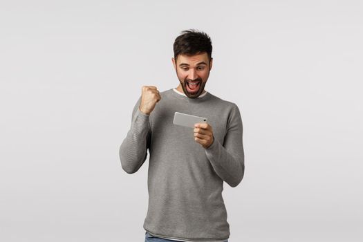 Happy and cheerful, excited bearded male in grey sweater, holding smartphone and fist pump as celebrating, say hooray or yes winning game level, see news in application, white background.