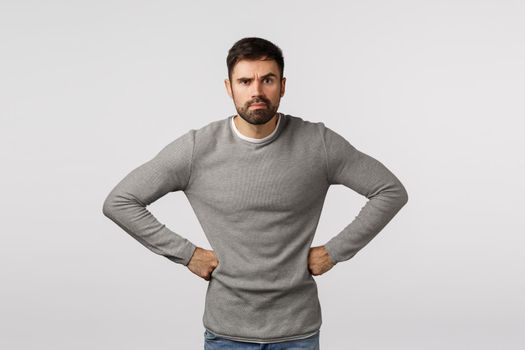 Angry and grumpy, displeased strict or serious male with beard, wear grey sweater, holding hands on waist in demanding, disappointed pose, frowning and grimacing upset, scolding someone.