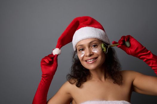 Cheerful pretty woman dressed as Santa uses jade roller massager for lymphatic drainage facial massage, poses on gray background with copy space for christmas advertisement of beauty salons. Skin care