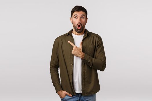 Excited and surprised, wondered handsome bearded male in coat seeing amazing opportunity, open mouth in amazement say wow, stare and pointing upper left corner thrilled, white background.
