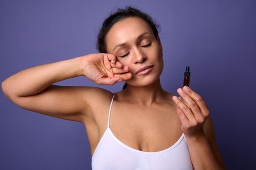 Attractive natural beauty African woman in white underwear holding jar with massage oil or smoothing anti-aging serum and posing against purple background with her eyes closed. Skin body care concept