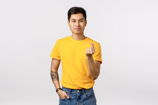 Cute asian hipster guy in yellow t-shirt, showing korean heart gesture and smiling, silly confess love, express affection or admiration, standing upbeat white background. Relationship concept.