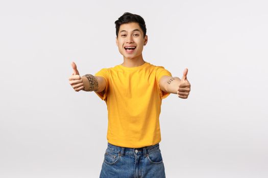 Good job, your doing great. Cheerful handsome asian male friend encourage you keep going, showing thumbs-up in approval, like product, recommend, agree with someone, standing white background.