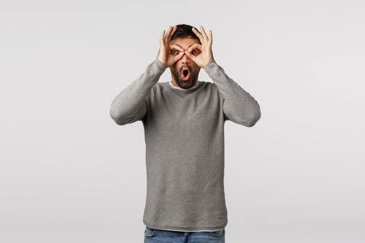 Surprised, excited and fascinated handsome bearded man in grey sweater, make circled with fingers over eyes like glasses or binocular, open mouth gasping amused, see and check out awesome promo.