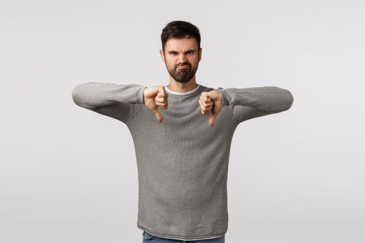 Rating, customer opinion and emotions concept. Angry and disappointed grumpy, bothered adult bearded man in grey sweater, grimacing with hateful expression, show thumbs-down, dislike gesture.