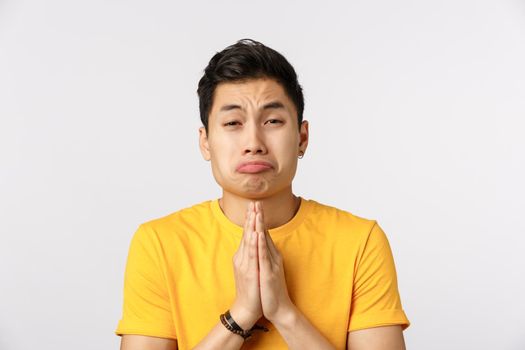 Clingy friend begging for favour with silly puppy look and pouting expression. Asian young man press hands together in pray, squinting asking mercy, say please, asking help, white background.