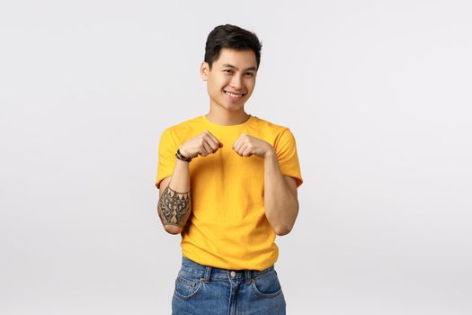 Silly and cute queer asian guy with tattoos acting all tender and innocent, making bunny dance with clenched fists near chest, smiling and laughing having fun, parying over white background.
