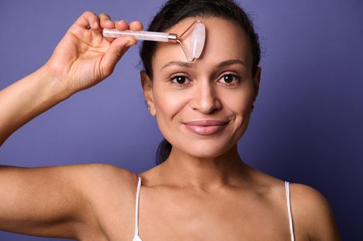 Beautiful middle aged woman with healthy perfect clean shiny skin and natural makeup massaging her face with pink stone jade roller massager, smiles looking at camera, isolated over violet background