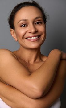 Close-up beauty portrait of self-confident mature African woman with perfect clean shiny healthy skin, hugging her self, smiling looking at camera against gray background.