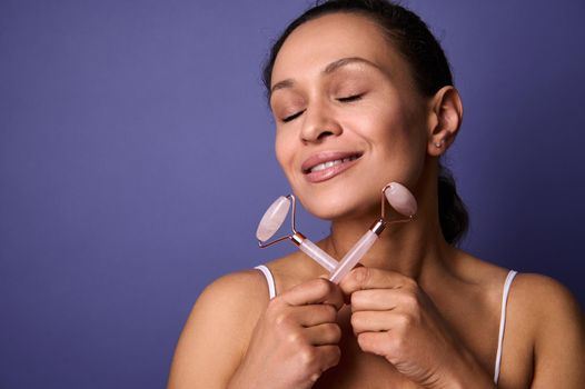 Relaxed middle aged 50-40 years old pretty woman posing with closed eyes, cute smiles, holding pink jade stone roller massagers in her hands, isolated over purple background with copy ad space