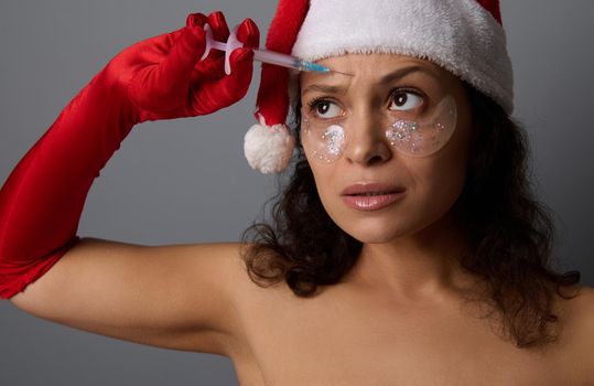 Anti-aging treatment, rejuvenation concept for Christmas advertising for spa salons, Beautiful woman wearing Santa hat and red gloves holds small syringe and makes beauty injection on her forehead