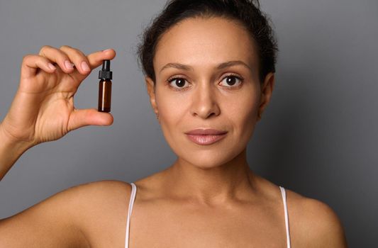 Beauty portrait of woman with perfect fresh clean skin looks at camera posing against gray background holding a bottle with beauty product. SPA, cosmetology, anti-aging therapy concept with copy space