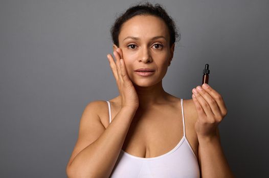 Beautiful middle aged woman holds a jar with beauty product, applies it on her face and performs anti-aging lymhpatic drainage massage for rejuvenating and skin smoothing. SPA, cosmetology concept