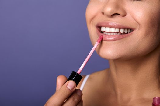 Close-up. Cropped view of attractive smiling woman with perfect clean healthy skin applying lip gloss hygienic balm on plump lips isolated on purple background. Copy space for ads. Make-up concept