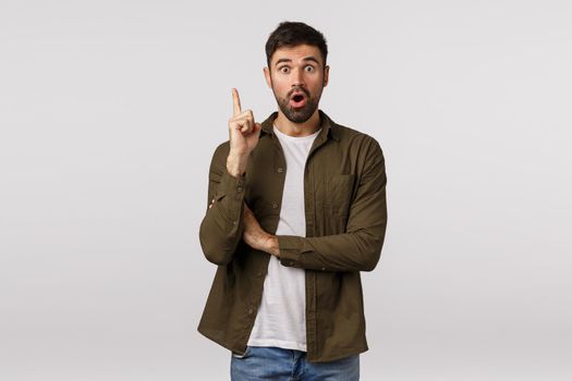 Got excellent idea, hear me out. Excited and striked young bearded man found solution, have interesting suggestion or resolve, raise index finger eureka gesture, gasping, say plan, white background.