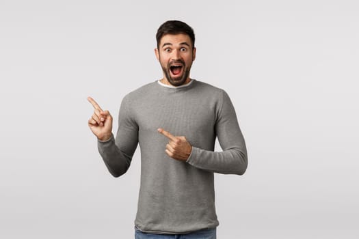 Excited, pleased and surprised handsome bearded adult man in grey sweater, smiling amused, showing pleasant good news, promo advertisement, pointing upper left corner, white background.