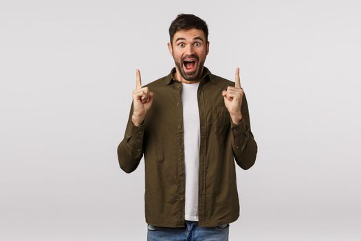 Guy adores discounts christmas time. Cheerful good-looking bearded male in coat, pointing fingers up and smiling excited, visit great event, feeling enthusiastic and thrilled, white background.