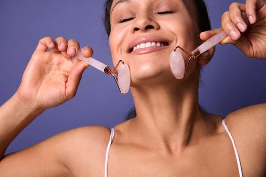 Close-up beauty portrait of a pretty woman with toothy smile using roller massager upon her face, doing lifting smoothing anti-aging lymphatic drainage facial massage. Violet background copy ad space