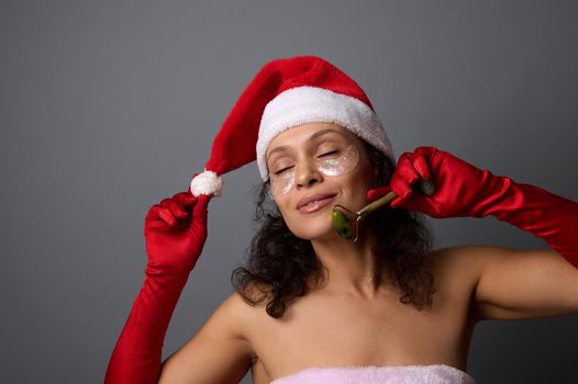 Attractive woman in Santa carnivaL attire uses jade roller massager for facelift and lymphatic drainage facial massage. Skin care, cosmetology concept for christmas advertisement of beauty spa salons