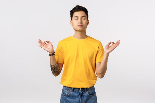 Patience, modern lifestyle concept. Calm and relieved young asian man taking emotions under control, close eyes breathing deep, make zen, mudra or nirvana sign, practice yoga, meditating.