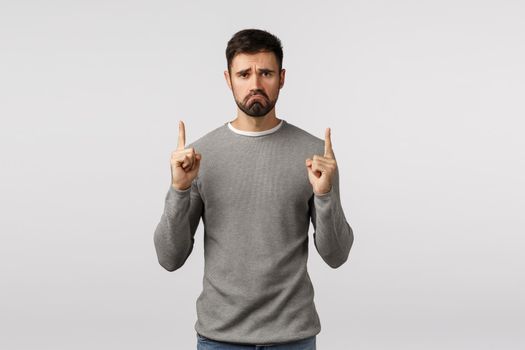 Miserable, upset and unsatisfied grumpy caucasian bearded guy, wear grey sweater, pointing up upset and distressed, feel regret or complain bad event happened, standing disappointed white background.