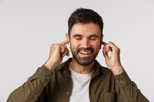 Cheerful and carefree, happy good-looking bearded male in coat, close eyes and smiling delighted, touching wireless earphones plugged in ears as enjoying nice sound quality, listen favorite music.