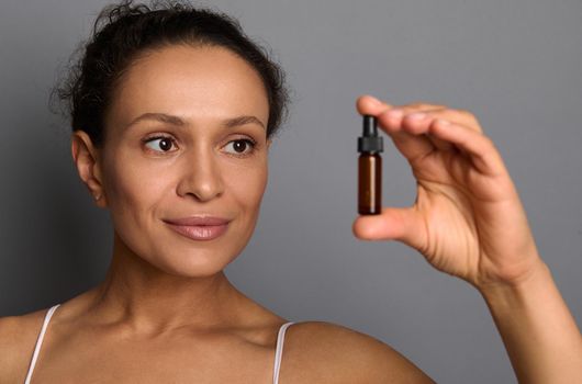 Close-up beauty portrait of beautiful woman with perfect fresh clean skin poses over gray background holding a bottle with beauty product. SPA, cosmetology, beauty treatment concept with copy space