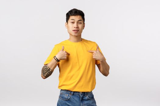 Ambitious handsome asian male snob in yellow t-shirt, with tattoos, pointing himself with confident, macho expression, praising own abilities, bragging how cool and awesome he is, white background.