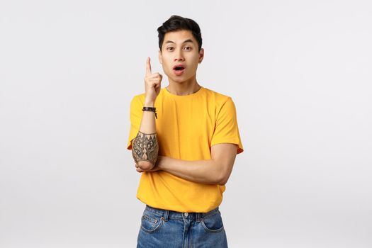 I have an assumption. Creative handsome asian male coworker, team member in yellow t-shirt, got plan, suggest way solution, raising index finger, standing opened mouth, saying what he think.