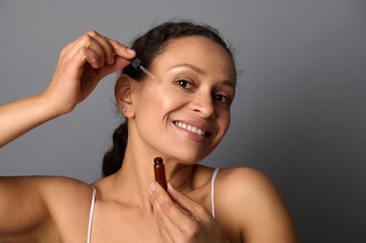 Charming middle aged woman smiles toothy smile applying a drop of cosmetic product on her face with dropper, isolated on gray background. Anti-aging, rejuvenation, hydration, body skin care concept