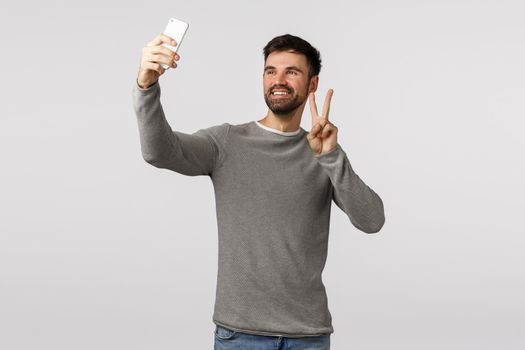 Optimistic and carefree, happy attractive male with beard in grey sweater, holding smartphone upwards as taking selfie, make peace sign and smiling, calling friend video-call online, white background.