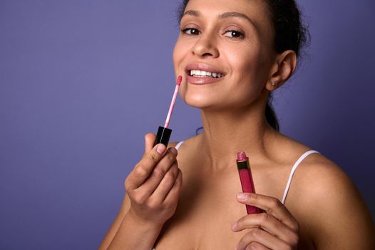 Charming natural beauty African woman applies lipstick, hygienic lip gloss to plump lips, smiles toothy smile looking at camer, posing over purple background with copy space for ads. Make-up concept