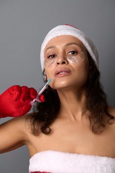 Gorgeous woman in Santa costume and smoothing patches under eye, holds syringe with beauty injection. Skin care rejuvenation, anti-aging care. Studio shot for beauty spa salons for Christmas giveaways