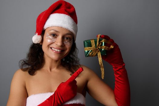 Cheerful African woman wearing cosmetic patches under eyes, and Santa outfit points with finger on Christmas gift box in shiny green wrapping paper and golden bow, smiles looking at camera. Copy space