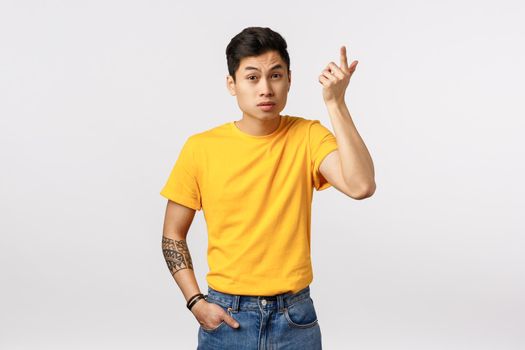 Wtf going on. Annoyed asian young man in yellow t-shirt, with tattoos, raising index finger in dismay and irritation, raising eyebrows as hearing nonsense, weird conversation, white background.