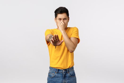 Eeew stinky. Disgusted asian male in yellow t-shirt, cover nose from aversion, smell something awful, pointing camera blame or accuse person reek, standing white background displeased.