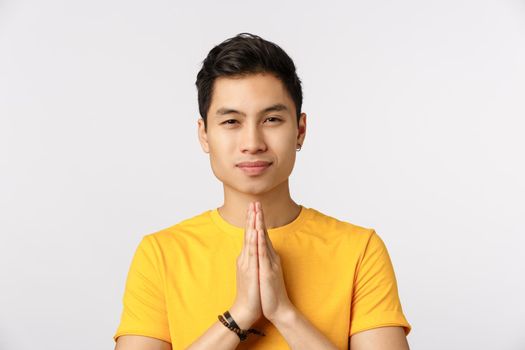 Customs, people concept. Attractive young chinese guy in yellow t-shirt, clasp hands together in pray, smiling joyfully, bowing politely, thanking, say namaste, standing white background.