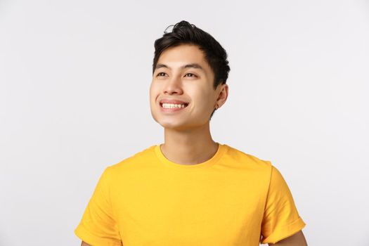 Proud and dreamy good-looking confident and cheerful asian man, wear yellow t-shirt, smiling delighted and contemplate something beautiful at upper left corner, white background. Copy space