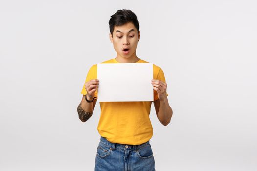 You need check this out. Astonished and impressed attractive young asian man in yellow t-shirt, holding blank piece paper and staring at sign with surprise and amazement, white background.