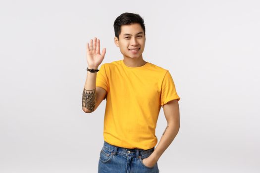 Friendly asian male student getting know new roommate in dorm, raising hand waving arm in hello, hi gesture, greeting friend, standing outgoing over white background, casually meeting someone.
