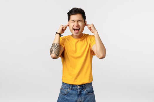 Depressed and tensed, young asian guy with tattoos, screaming from pain or agony, fed up, close eyes and ears with fingers, annoyed loud bothered noise, siren, standing displeased white background.