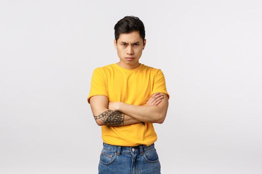 I am mad at you. Angry and displeased, offended defensive asian man in yellow t-shirt, cross arms over chest, frowning sulking and look from under forehead insulted, white background.