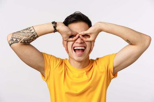 Fun, excitement and enthusiasm concept. Cheerful and carefree playful asian tattooed guy in yellow t-shirt, making glasses or superhero mask with fingers over eyes, standing white background joyful.