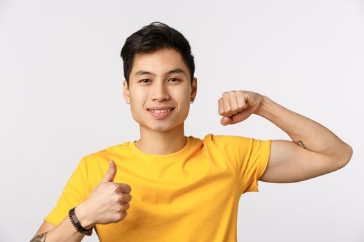 Time pump it up. Attractive young asian man in yellow t-shirt showing muscle and thumb-up, smiling, encourage do physical exercises, come gym together, want gain strong biceps.