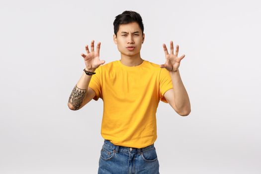 Roar hot staff. Cheeky and handsome stylish asian man in yellow t-shirt, making paw gesture as if squeezing something or pretend tiger, squinting sassy and standing white background.
